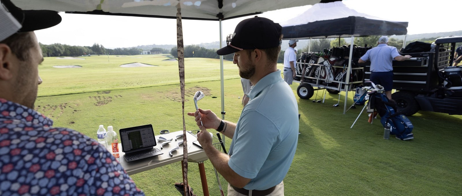 A Next Gear fitter talking a customer through an iron head beside the launch monitor data on the fitting cart